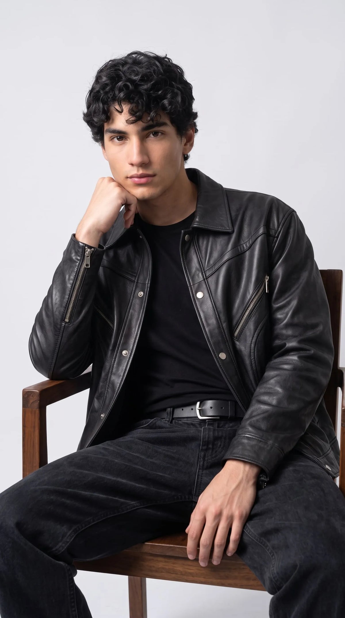 Contemporary men's fashion featuring a darkest-black genuine leather jacket with oversized sleeves, angled silver chest zippers and a snap-button front, worn over a black T-shirt with dark charcoal jeans. The relaxed seated pose highlights the loose silhouette and textured leather surface, creating an editorial street-style outfit that blends everyday comfort with confident, understated luxury.