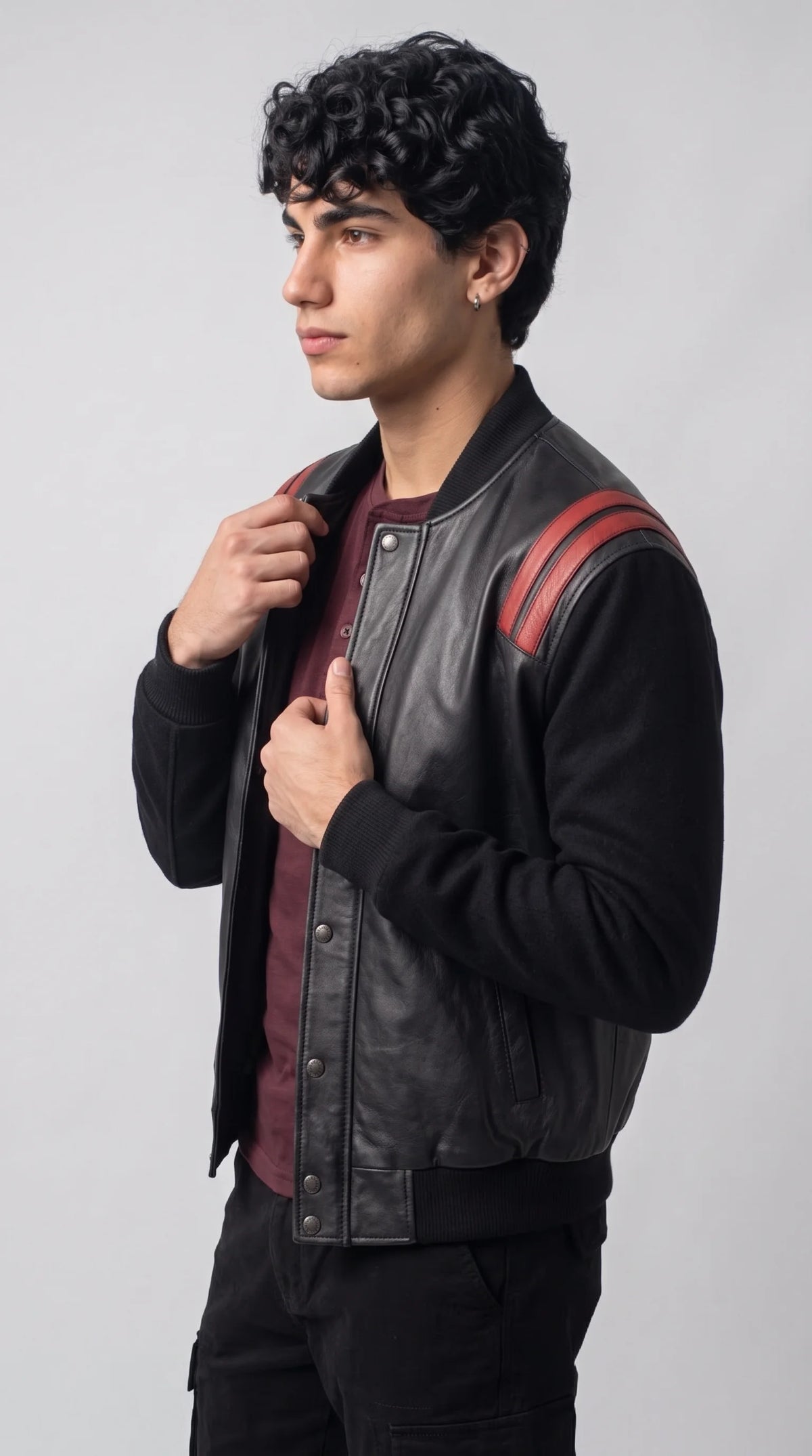 Men’s black and red leather varsity jacket styled over a burgundy Henley with dark cargo trousers for a sharp modern-city outfit. Red shoulder stripes stand out against the black leather front and textured sleeves, while the snap-button closure and ribbed trim preserve its athletic bomber character. A versatile statement layer for casual evenings and contemporary urban wardrobes.