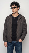 Men’s dark charcoal-grey silk-cotton hooded jacket styled over a black button-neck Henley shirt with dark blue jeans and slim black sunglasses. The adjustable drawstring hood, full-length metal zipper and softly textured fabric create a relaxed urban look, ideal for modern streetwear, weekend layering and understated everyday fashion.