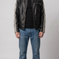 A man wearing a black leather motorcycle jacket with white stripes on the sleeves, a black t-shirt, blue jeans, and black shoes, standing against a light gray studio background.