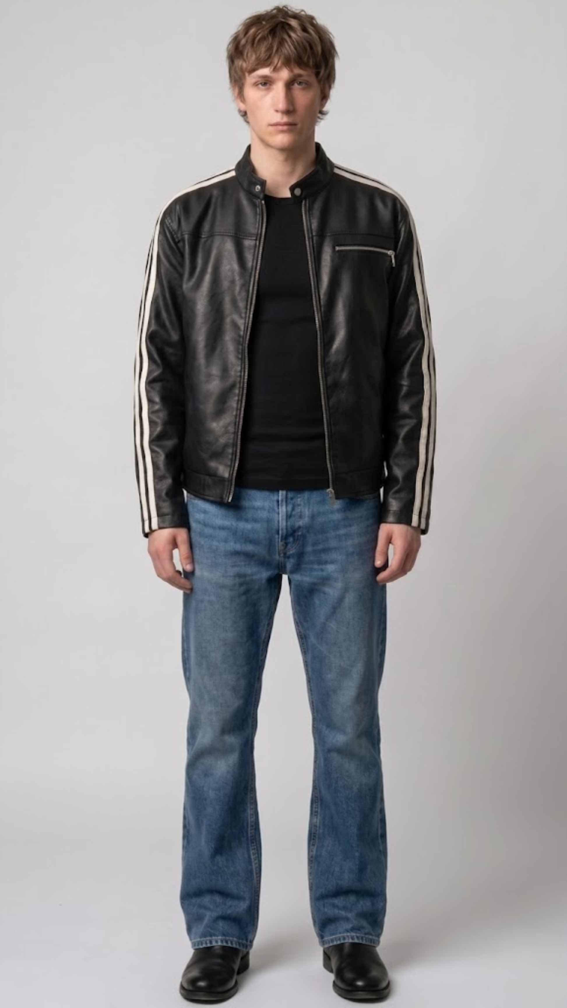 A man wearing a black leather motorcycle jacket with white stripes on the sleeves, a black t-shirt, blue jeans, and black shoes, standing against a light gray studio background.