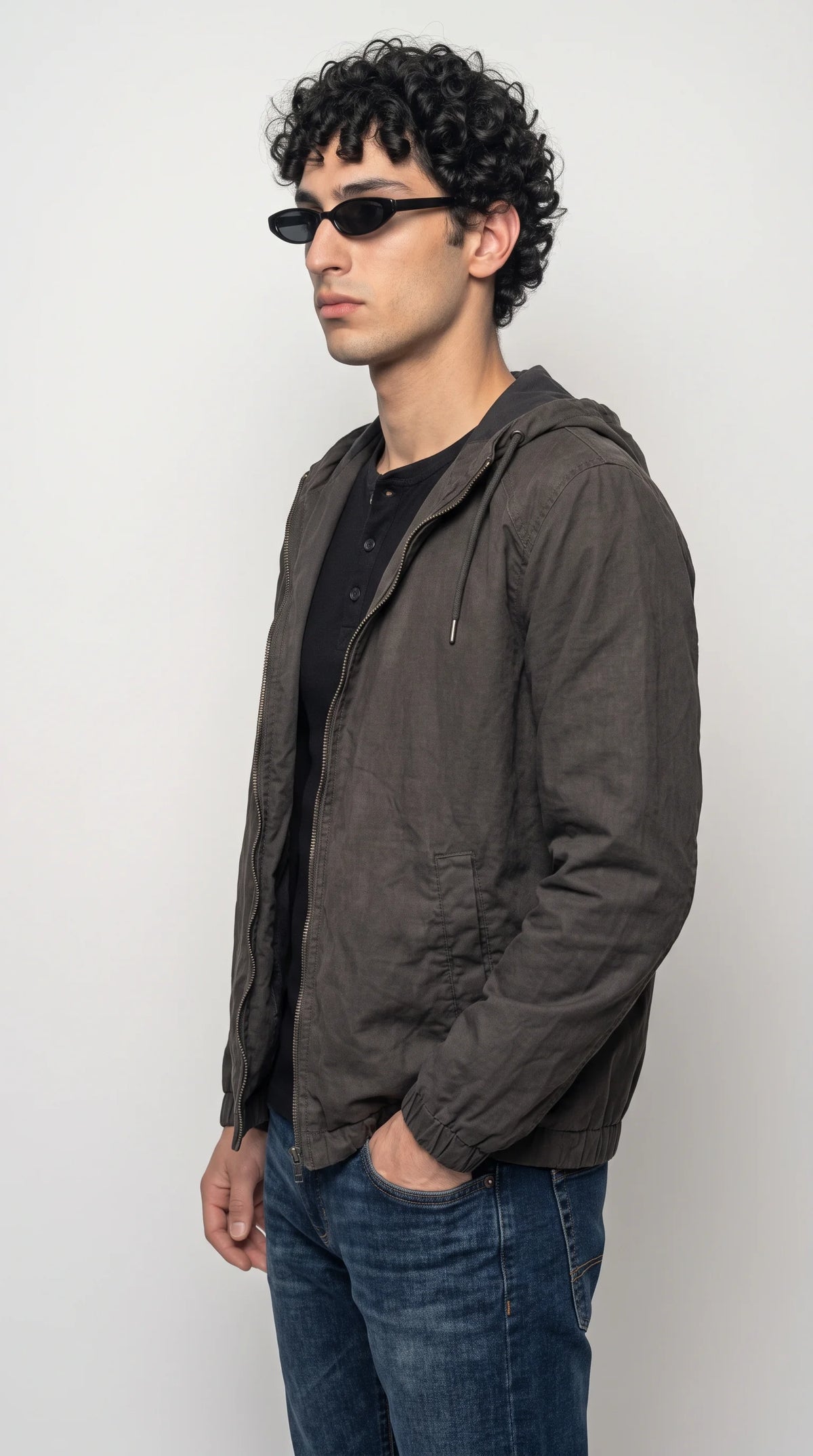 Men’s dark ash-grey hooded zip jacket with discreet side waist pockets, elasticated cuffs, an adjustable hood and a streamlined silhouette, worn with a black top and blue jeans. The subtle fabric texture and neutral colour make it a versatile piece for modern city wear, travel outfits and understated streetwear without oversized graphics or loud detailing.