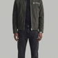 A man modeling a dark olive green suede jacket over a black shirt and dark gray jeans, standing against a solid light gray background.