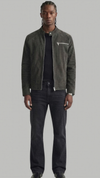 A man modeling a dark olive green suede jacket over a black shirt and dark gray jeans, standing against a solid light gray background.