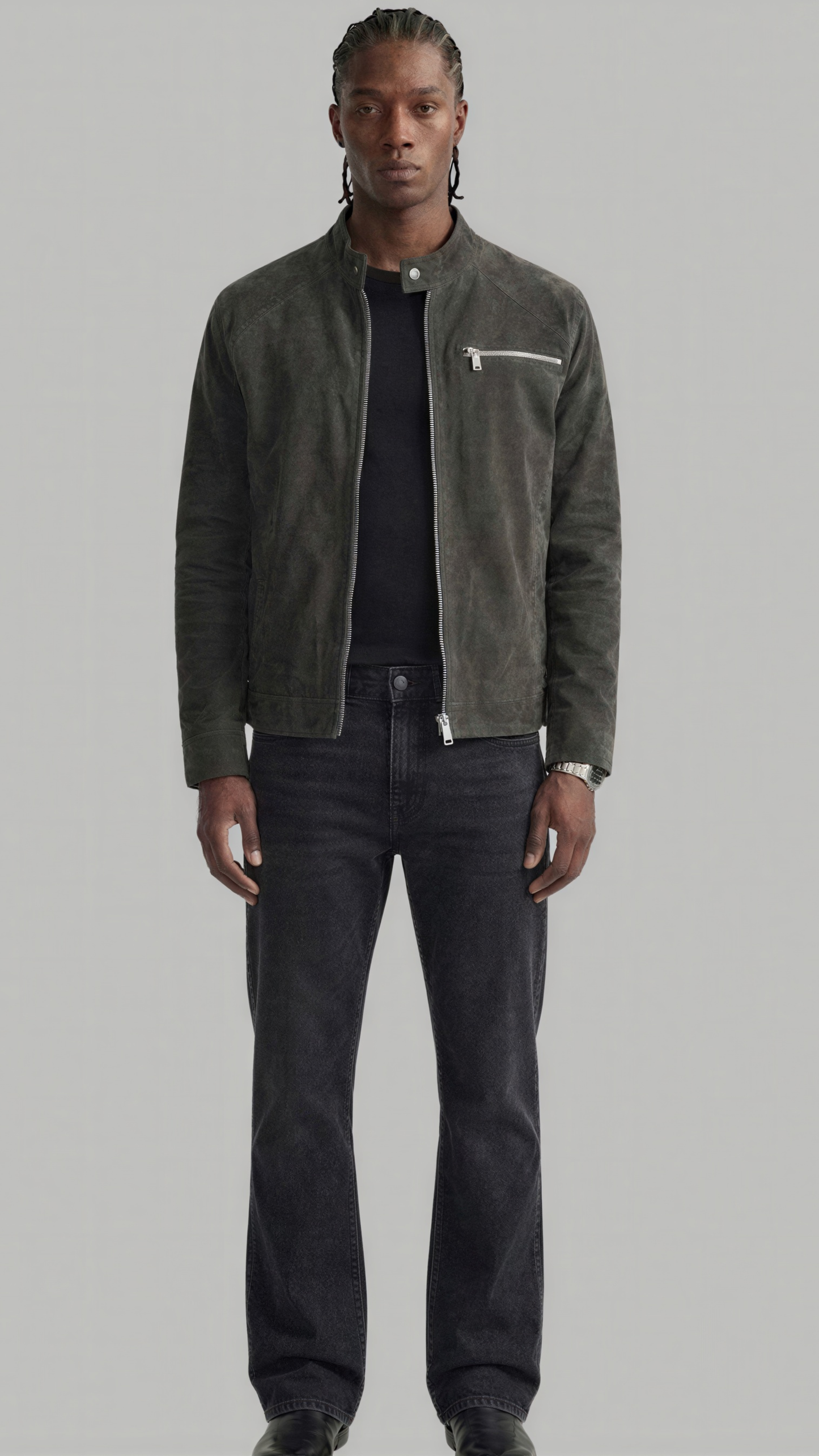A man modeling a dark olive green suede jacket over a black shirt and dark gray jeans, standing against a solid light gray background.