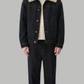 A model stands against a solid gray background wearing a dark indigo denim jacket with a beige shearling-style collar, a cream t-shirt, dark denim pants, and black shoes.