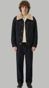 A model stands against a solid gray background wearing a dark indigo denim jacket with a beige shearling-style collar, a cream t-shirt, dark denim pants, and black shoes.