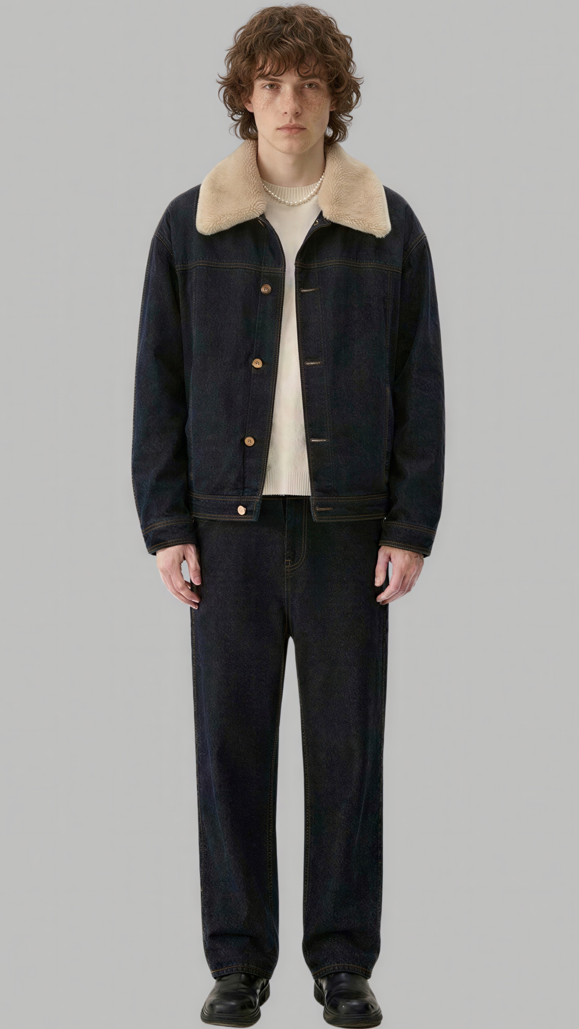 A model stands against a solid gray background wearing a dark indigo denim jacket with a beige shearling-style collar, a cream t-shirt, dark denim pants, and black shoes.