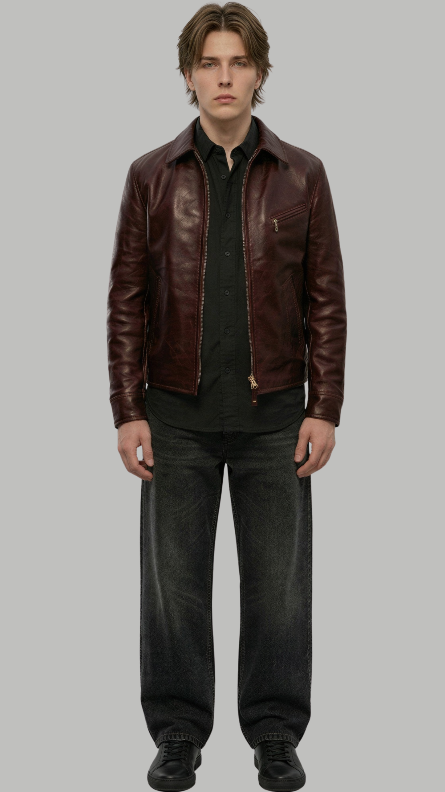 A male model wearing a dark brown leather jacket over a black shirt and dark gray jeans, standing against a solid gray background.