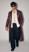 Men’s dark wine-red genuine leather long coat styled with an open ivory shirt, textured mesh vest, wide black trousers, chunky black shoes and dark sunglasses. The rich burgundy finish, classic notch lapels and elongated silhouette create a confident luxury editorial look that blends contemporary menswear, refined evening styling and bold urban fashion.