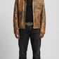 A man modeling a distressed brown leather jacket over a black shirt and dark jeans, standing against a light gray studio background.