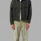 A man modeling a dark washed denim jacket over a white t-shirt, paired with light olive green pants and white sneakers, against a solid gray studio background.