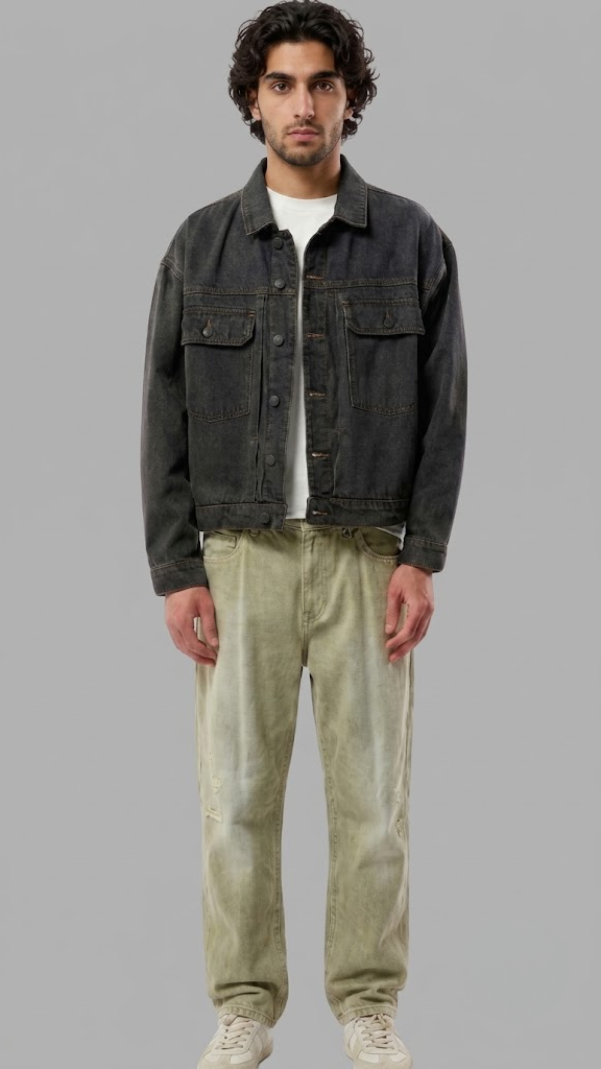 A man modeling a dark washed denim jacket over a white t-shirt, paired with light olive green pants and white sneakers, against a solid gray studio background.