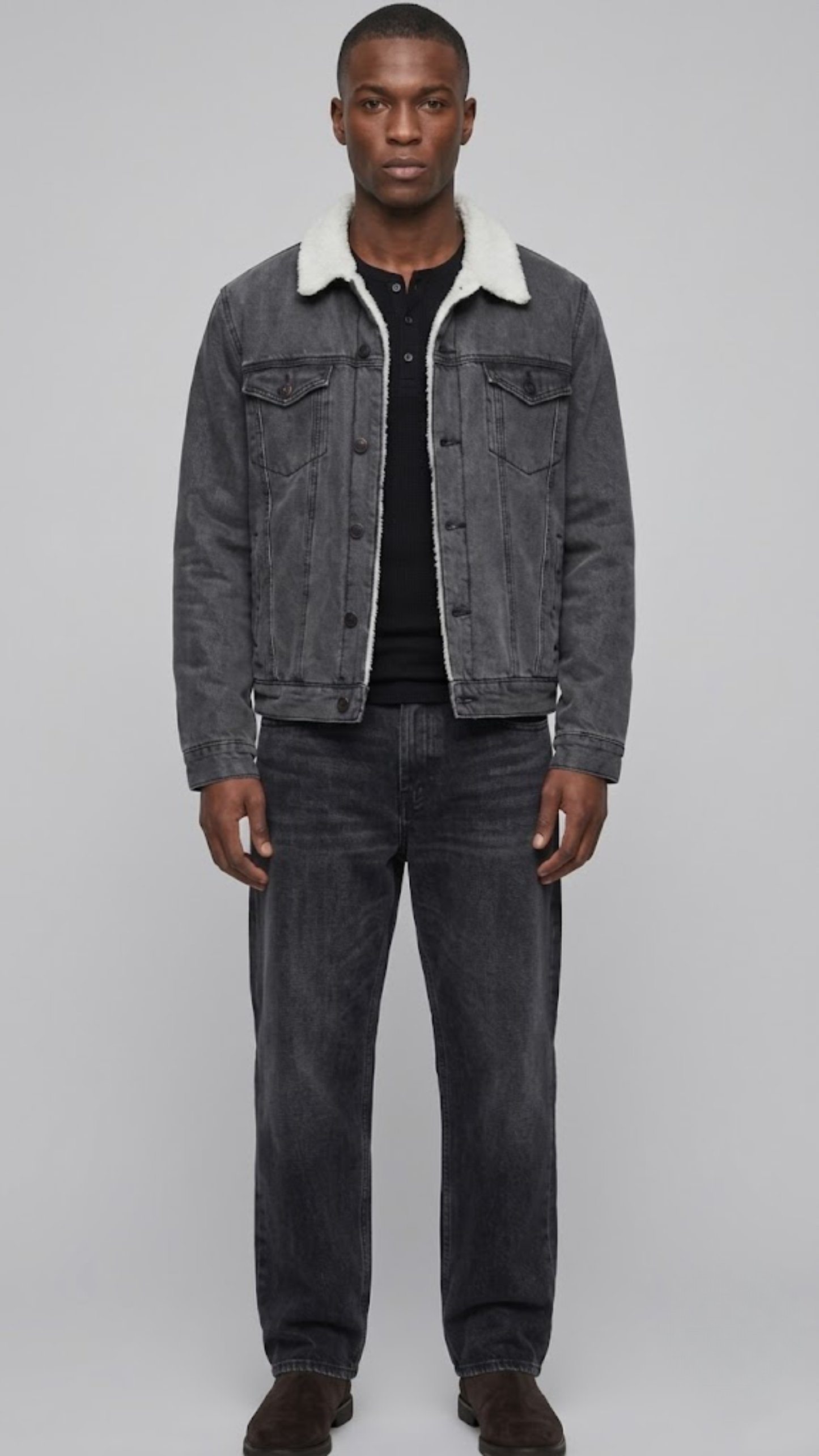 A man modeling a dark gray washed denim jacket with a white sherpa collar, worn over a black t-shirt and dark gray jeans, against a solid light gray background.