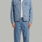 A man modeling a light blue denim jacket and matching light blue denim jeans over a white t-shirt, standing against a solid gray studio background.