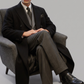 Man in a black double breasted long wool coat sitting on a gray chair against a plain background