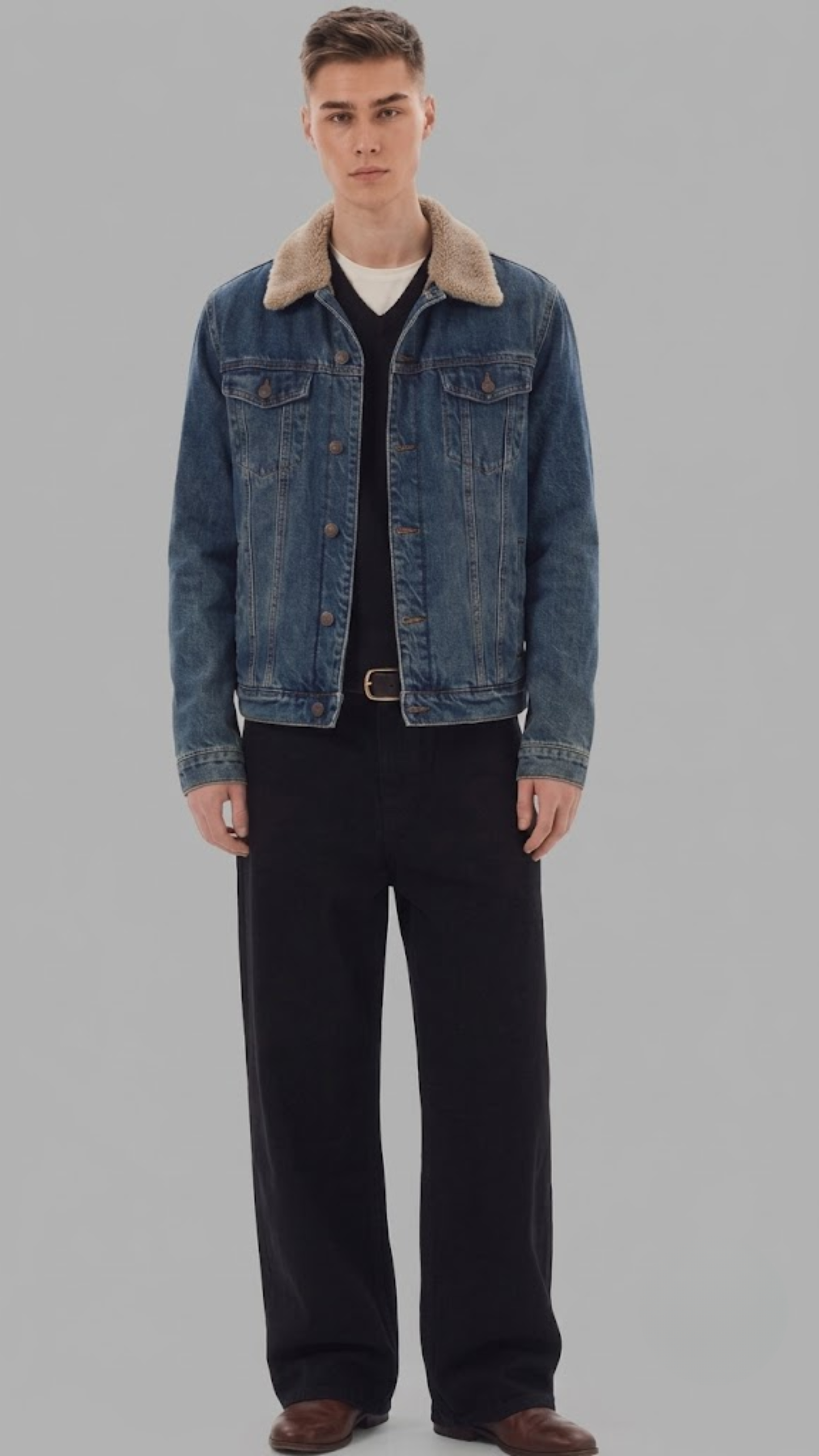 A man modeling a blue denim jacket with a beige shearling-style collar, worn open over a black shirt and dark trousers, against a solid gray studio background.