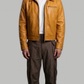 Man wearing a tan leather jacket and brown pants on a gray background
