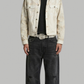 A model stands against a solid gray background wearing a light beige distressed denim jacket over a black t-shirt, black baggy jeans, and silver sneakers.