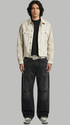 A model stands against a solid gray background wearing a light beige distressed denim jacket over a black t-shirt, black baggy jeans, and silver sneakers.