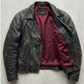 A distressed black leather motorcycle jacket with a deep burgundy red lining, laid flat on a textured gray concrete surface.