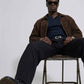 men wearing a jackero brown jacket, a dark blue polo shirt, and black trousers sits on a metal stool against a plain white studio background.
