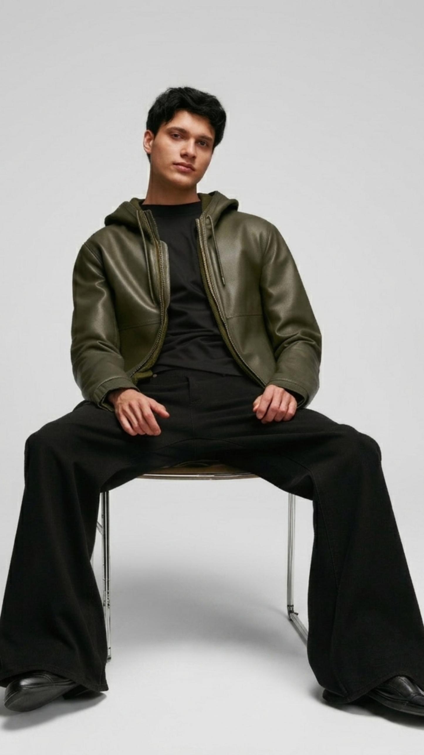 Man sitting on a chair wearing a green leather jacket and black pants against a white background