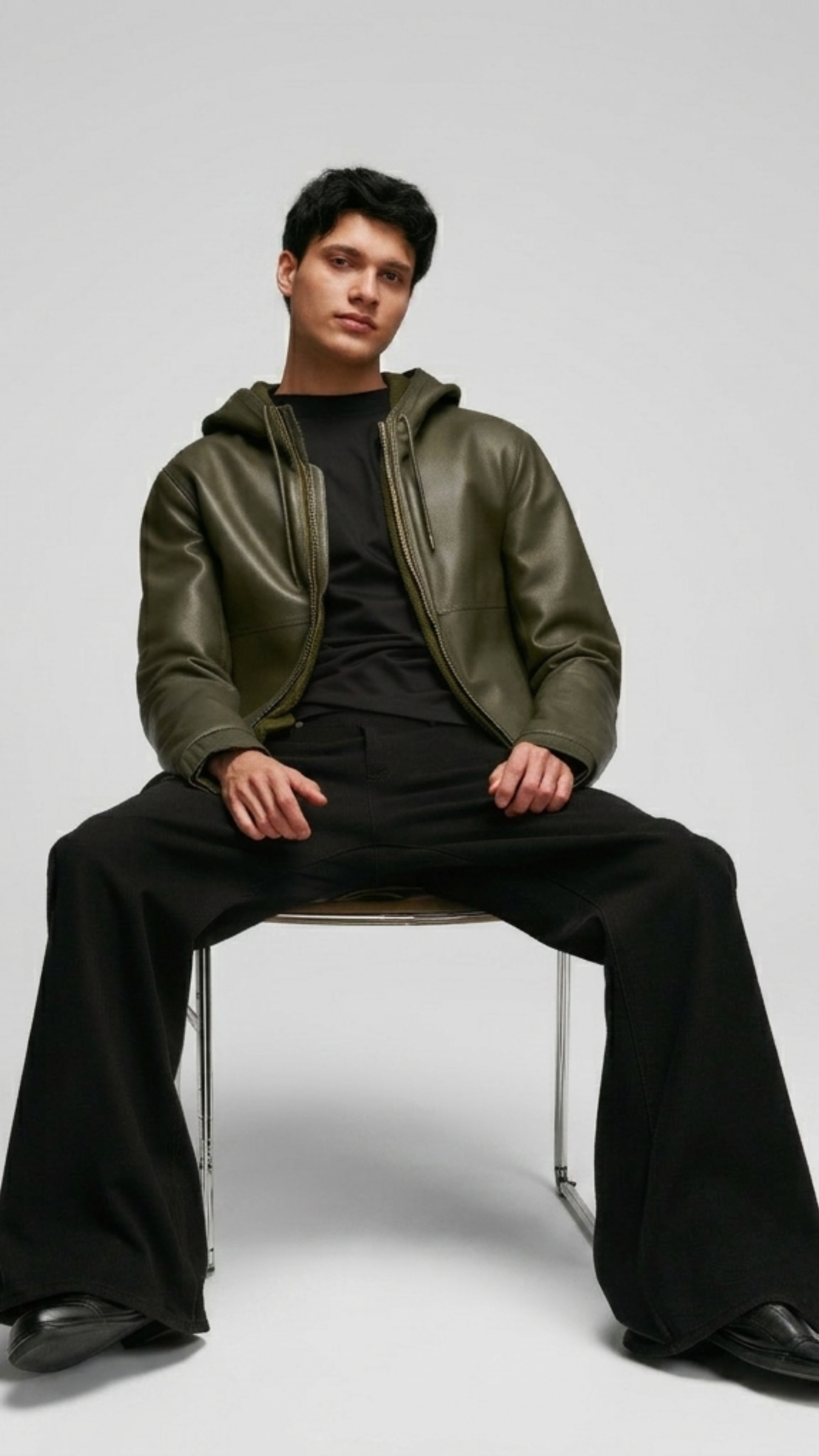 Man sitting on a chair wearing a green leather jacket and black pants against a white background
