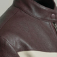 Close-up of a dark brown leather jacket with a high collar and a horizontal cream stripe across the chest.