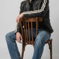 A man wearing a black leather jacket with cream stripes on the sleeves, blue jeans, and black boots, sitting on a wooden chair against a light gray studio background.