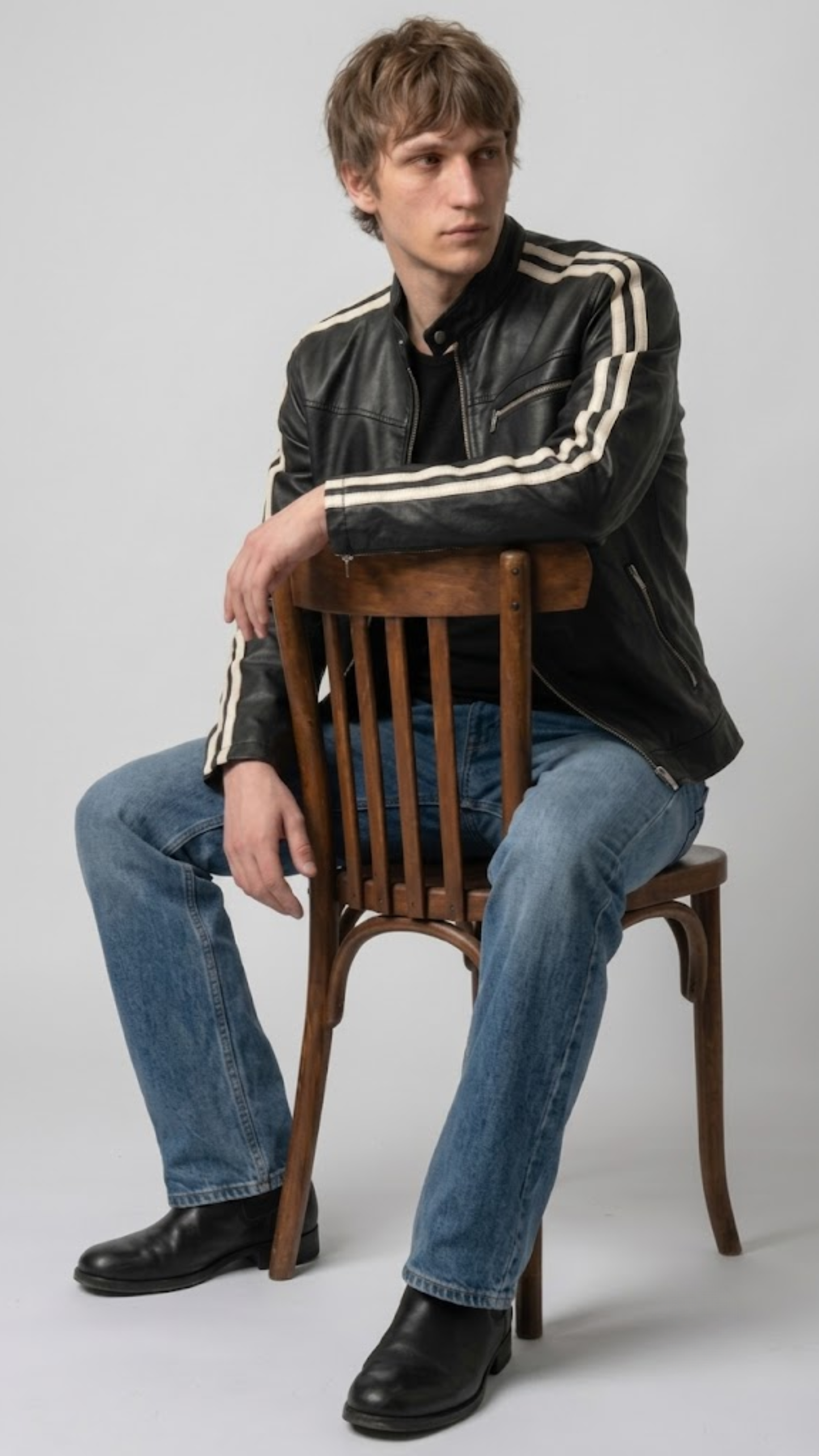 A man wearing a black leather jacket with cream stripes on the sleeves, blue jeans, and black boots, sitting on a wooden chair against a light gray studio background.