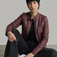 A woman with curly hair sits on a modern chair wearing a dark brown leather jacket with cream stripes, dark blue jeans, and white sneakers against a light gray studio background.