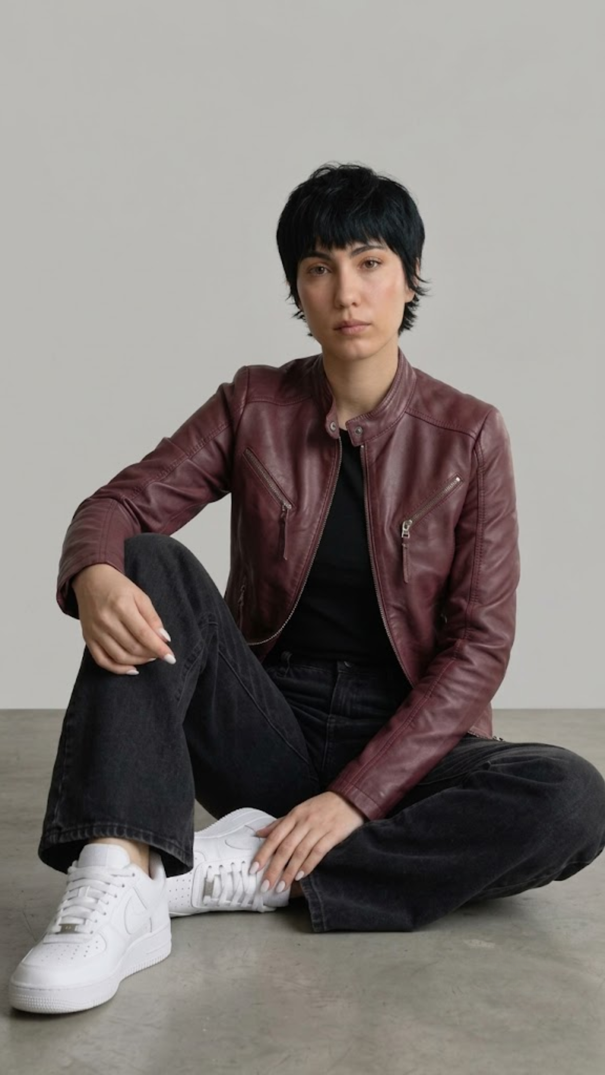 A woman with curly hair sits on a modern chair wearing a dark brown leather jacket with cream stripes, dark blue jeans, and white sneakers against a light gray studio background.