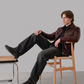 Man sitting on a chair with one leg crossed over the other, wearing a brown leather jacket and dark pants.