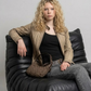 A women wearing a beige leather jacket over a white t-shirt and brown pants, sitting on a concrete block against a gray studio background.