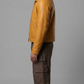 A man in profile wearing a mustard yellow leather jacket, brown cargo pants, and brown shoes, standing against a solid gray studio background.