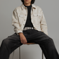 A man with curly dark hair sits on a metal chair wearing a light beige denim jacket over a black t-shirt and black jeans against a solid gray studio background.