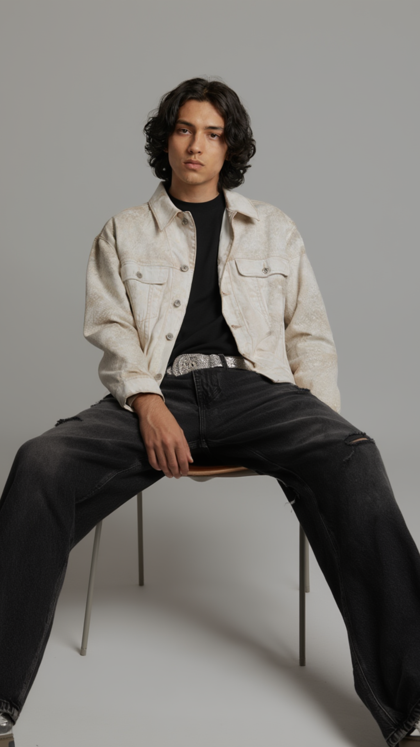 A man with curly dark hair sits on a metal chair wearing a light beige denim jacket over a black t-shirt and black jeans against a solid gray studio background.