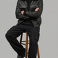Man sitting on a stool wearing a black leather jacket with black shearling and pants against a gray background