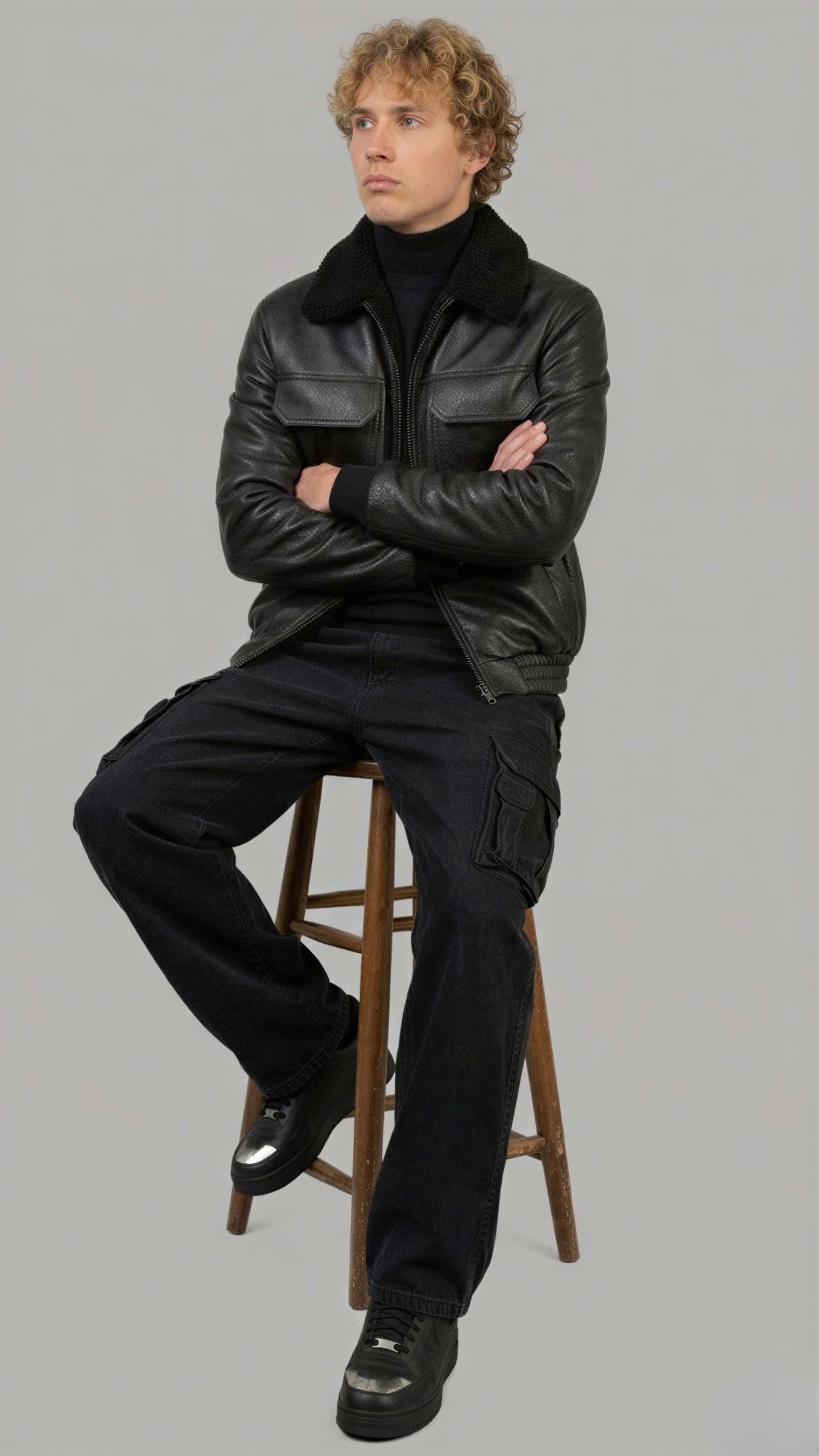 Man sitting on a stool wearing a black leather jacket with black shearling and pants against a gray background