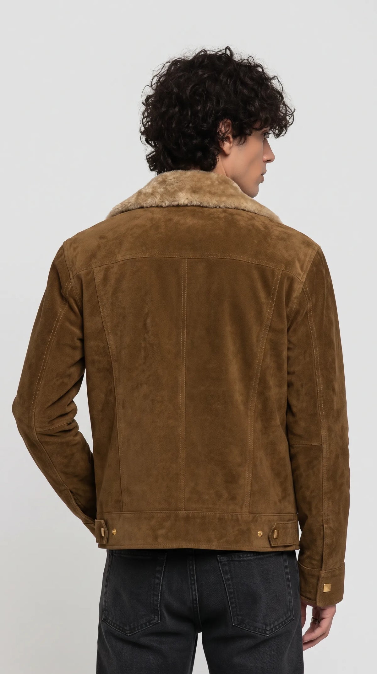 Dark mustard-brown genuine suede Western trucker jacket featuring a neatly stitched upper-back yoke, long vertical panel seams and adjustable hem tabs with metal fasteners. The plush light-brown faux-shearling collar contrasts with the matte suede surface, while the structured construction gives this heritage-inspired outerwear a rugged, refined appearance suited to dark jeans, boots and understated workwear outfits.