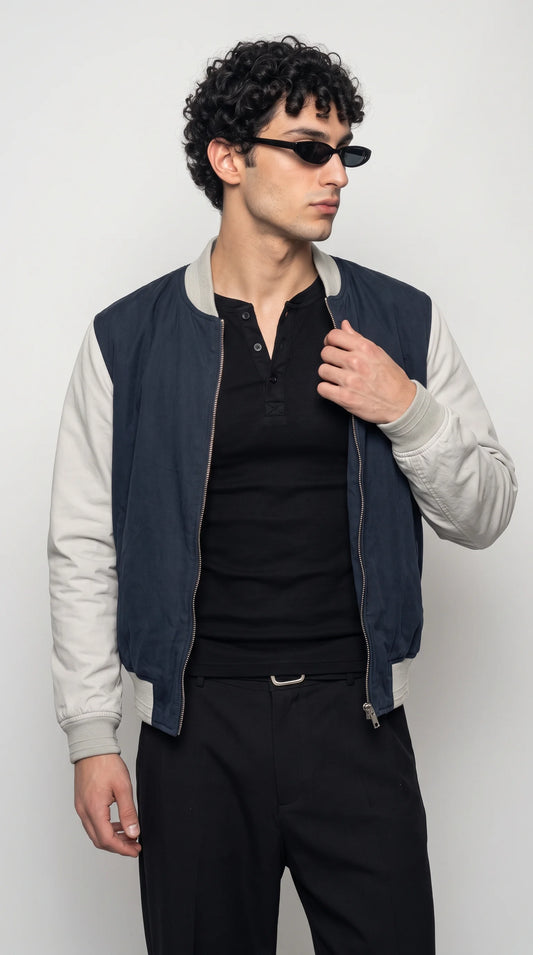 Men’s dark navy-blue cotton bomber jacket with contrasting light-grey sleeves, a ribbed collar, elasticated cuffs and hem, and a full-length metal zipper, styled with a black henley and black trousers for a clean varsity-inspired streetwear outfit.