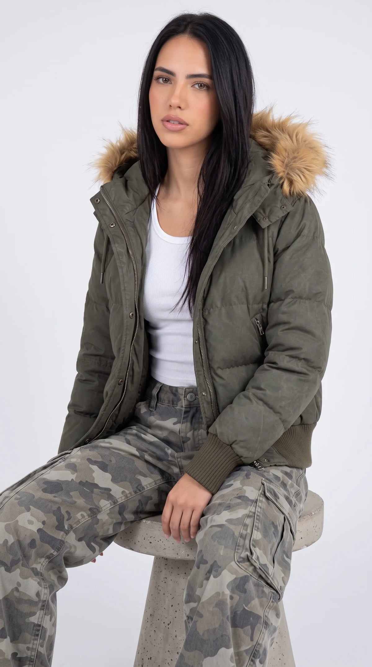 Relaxed women's winter outfit featuring an olive-green oversized puffer jacket with a plush tan fur-trimmed hood, worn over a white ribbed tank and grey camouflage cargo jeans while seated on a minimalist studio stool. The contrasting textures of the padded jacket, soft hood trim and utility trousers create a contemporary fashion-editorial look with relaxed proportions and an understated outdoor-inspired edge.