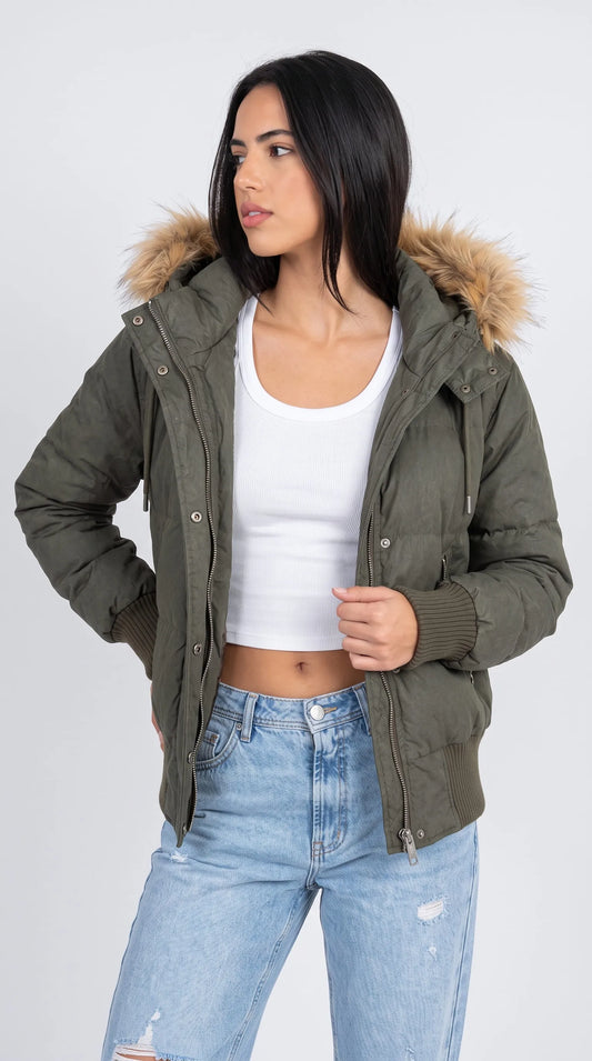 Women's olive-green oversized quilted puffer jacket with a voluminous tan faux-fur-trimmed hood, full-length metal zipper, snap-button front fastening and ribbed cuffs. Styled over a white fitted crop top with distressed light-wash blue jeans, this relaxed winter streetwear outfit combines a muted military-inspired colour palette with cosy texture and a modern cropped bomber silhouette.