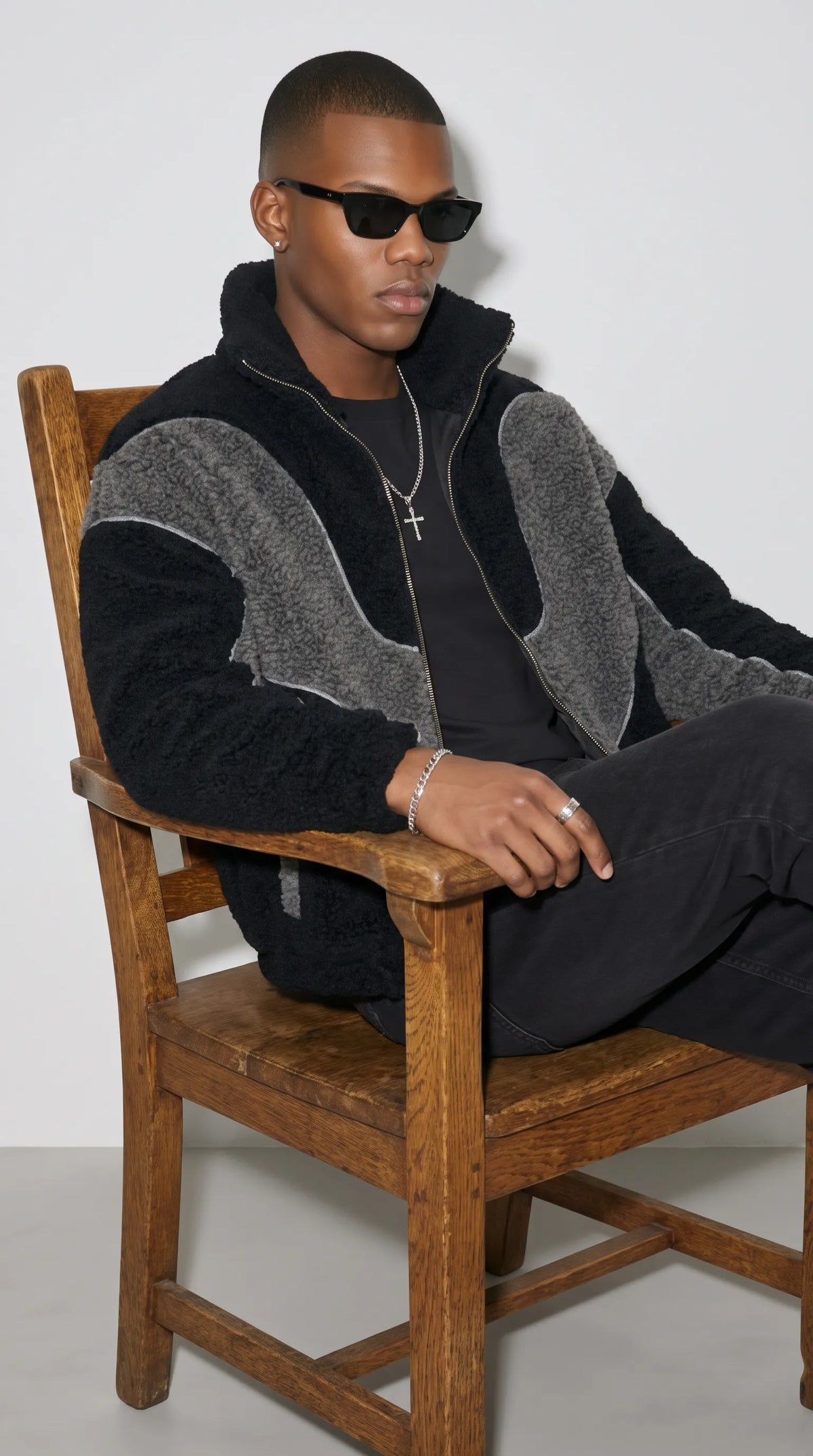 A modern men’s streetwear outfit featuring an oversized black and dark grey Y2K fleece jacket, styled with a black T-shirt, dark jeans, sunglasses and silver accessories while seated on a wooden chair. The plush colour-block panels and high collar create a strong visual statement, blending relaxed casual styling with a bold editorial look for cooler-weather city outfits.