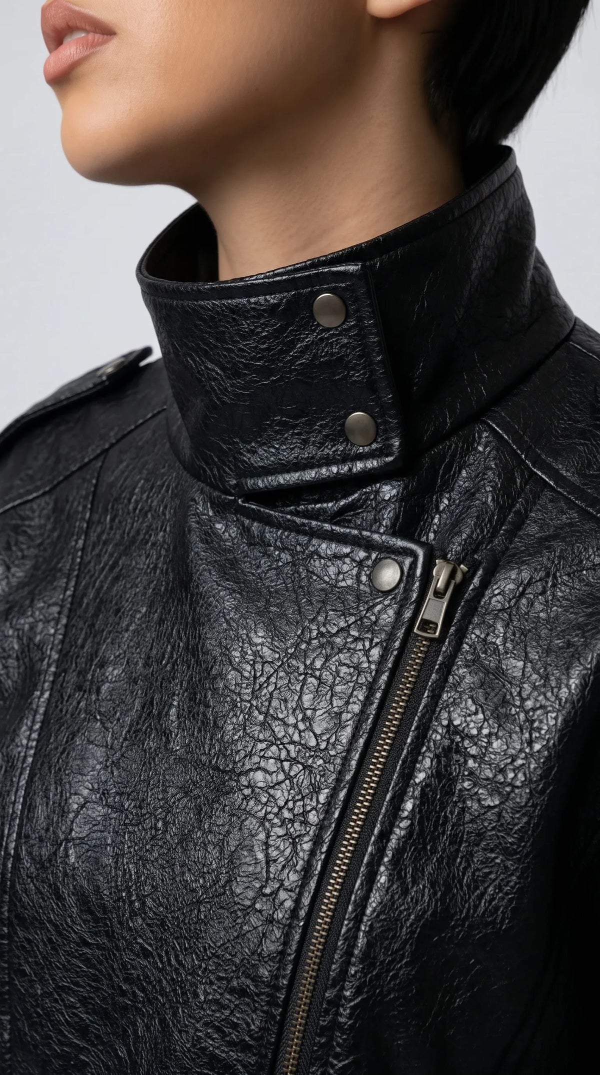 Close-up of a black crinkled genuine leather jacket showing its sculptural wrap-around high collar, metallic snap buttons, offset front zipper and distinctive textured finish for an edgy luxury outerwear aesthetic.