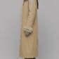 A model wearing a pastel yellow textured cotton long-coat, a black top, and cream wide-leg trousers, standing against a light gray studio background.