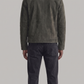 A man viewed from the back wearing a dark olive green suede jacket and dark charcoal gray jeans, standing against a solid light gray background.