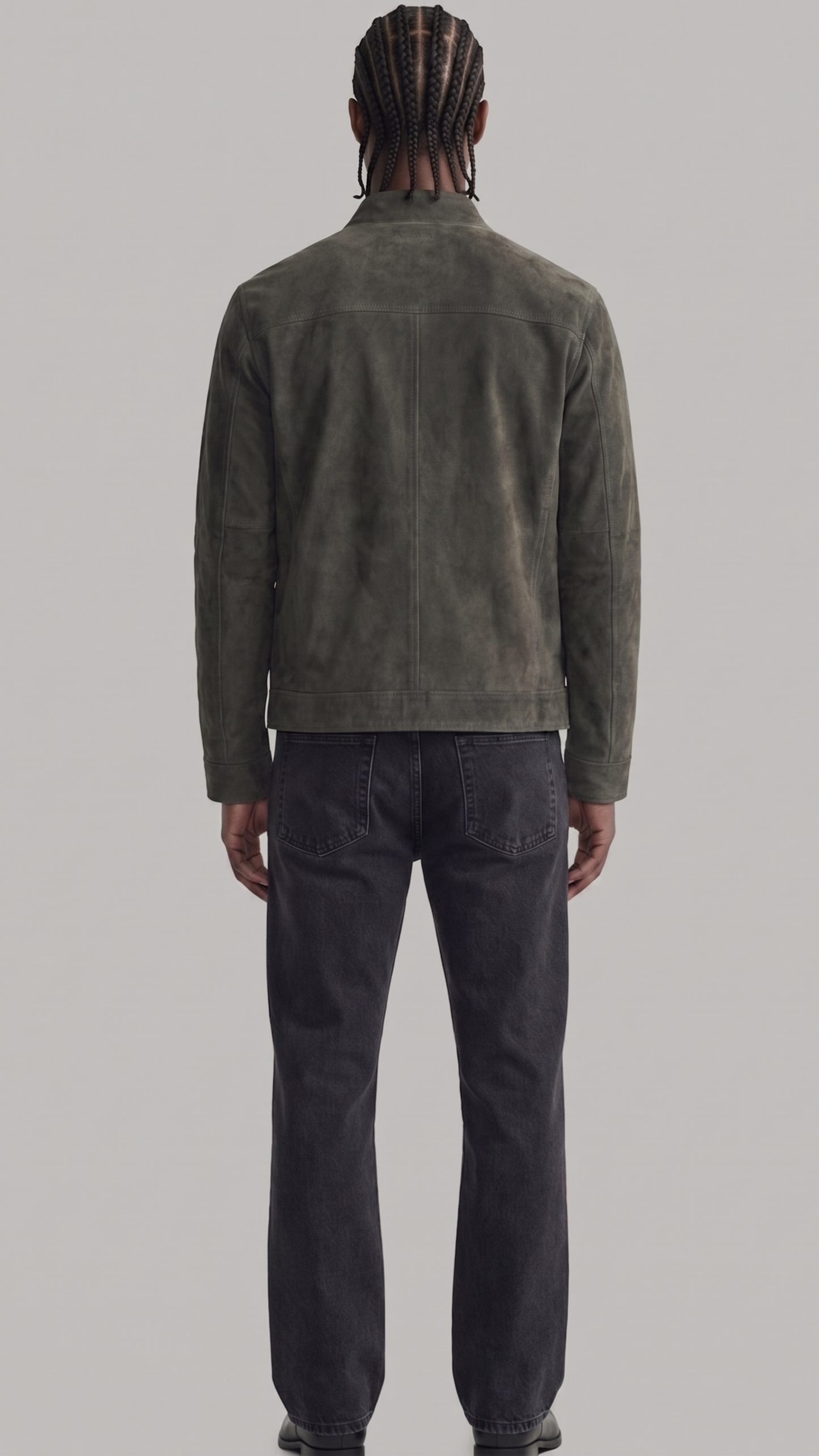 A man viewed from the back wearing a dark olive green suede jacket and dark charcoal gray jeans, standing against a solid light gray background.