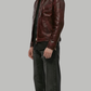 A man wearing a dark burgundy leather jacket, black shirt, black jeans, and black sneakers, standing in profile against a solid gray background.